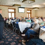A man stands in front of a room with each seat taken by a listener focused on him